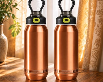 Op de afbeelding: Twee identieke koperkleurige metalen waterflessen met een cilindrisch lichaam en horizontale ribbels bij de basis. Elke fles heeft een zwarte schroefdop met een limoengroene knop en een zwarte ronde draagbeugel aan de bovenzijde.
