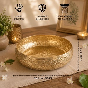 An empty gold metal urli bowl with embossed exterior detailing and an engraved starburst pattern on the inside base, showing product dimensions of 26.5 cm by 7 cm and feature icons.