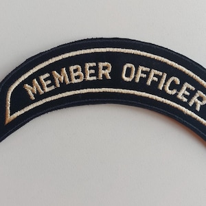Może przedstawiać: Zakrzywiona, czarna naszywka z wyhaftowanym napisem MEMBER OFFICER pogrubionymi, jasnymi wielkimi literami. Naszywka posiada pasującą jasną obwódkę, która podąża za łukowatym kształtem wzoru, tworząc schludny i profesjonalny wygląd.