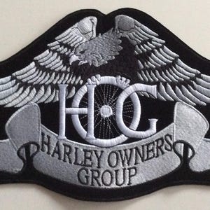 Könnte beinhalten: Ein schwarz-silberner Stickaufnäher mit einem Adler mit ausgebreiteten Flügeln über den Buchstaben HOG und einem Banner darunter mit dem Text HARLEY OWNERS GROUP. Das Design enthält ein eingetragenes Markenzeichen auf der rechten Adlerflügelseite.