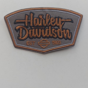 Peut inclure: Un écusson brodé en forme de bouclier avec un fond gris et une bordure cuivrée. Le texte indique Harley Davidson en lettres cuivrées stylisées avec des contours noirs, incluant l'inscription EST 1903 et un logo central en forme de barre et bouclier.