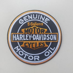 Peut inclure: Un écusson brodé circulaire avec un fond noir et une bordure dorée et blanche. Le texte indique Genuine, 5 Gallons, Motor, Harley-Davidson, Cycles et Motor Oil. L'emblème central est un logo en forme de barre et de bouclier noir et doré.