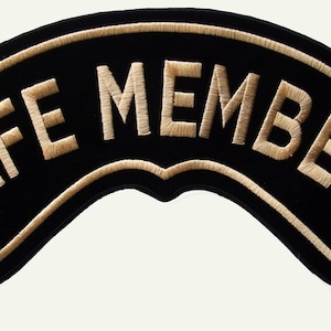 Puede incluir: Un parche bordado negro de forma curva que presenta el texto "LIFE MEMBER" cosido con hilo de color beige claro. El parche tiene un borde a juego en beige claro que sigue la forma arqueada del diseño, creando un contorno limpio.