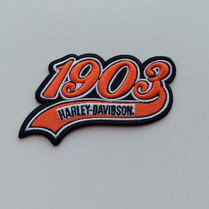 Puede incluir: Un parche bordado que presenta los números 1903 en color naranja con contorno blanco y negro. Debajo de los números, una banda naranja curva con borde blanco contiene el texto negro HARLEY-DAVIDSON, sobre un fondo negro sólido.