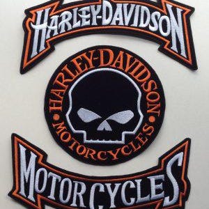 Op de afbeelding: Een set van drie geborduurde patches met het Harley-Davidson logo. De bovenste en onderste patches zijn gebogen banieren met witte tekst en oranje randen. De middelste patch is cirkelvormig met een schedel en bijbehorende tekst.