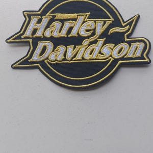 Może przedstawiać: Okrągła haftowana naszywka z napisem Harley Davidson wykonanym białymi literami ze złotymi obrysami. Wzór znajduje się na czarnym tle ze złotą okrągłą obwódką oraz ostrymi, kanciastymi złotymi akcentami rozciągającymi się po bokach.