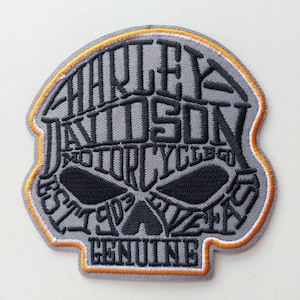 Può includere: Una toppa ricamata grigia a forma di teschio, con testo nero che recita Harley Davidson Motorcycles Est 1903 Genuine. Il design presenta un sottile bordo arancione lungo il perimetro, con cuciture nere che formano le orbite del teschio.