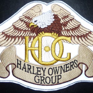 May include: An embroidered patch featuring a bald eagle with spread wings above a gold HOG logo and a tan banner with the text HARLEY OWNERS GROUP. The design includes a white border and a registered trademark symbol on a dark background.