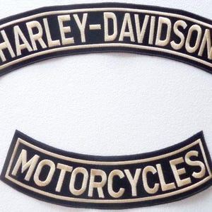 Op de afbeelding: Twee gebogen geborduurde patches met een zwarte achtergrond en crèmekleurige randen en letters. De bovenste patch bevat de tekst HARLEY-DAVIDSON, terwijl de onderste patch de tekst MOTORCYCLES bevat. Beide patches hebben een gestikte afwerking.