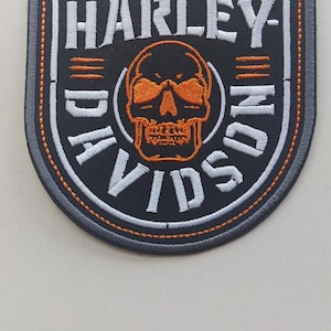 Può includere: Una toppa ricamata a forma di scudo con sfondo nero e un grafico centrale di un teschio arancione. Il testo HARLEY-DAVIDSON è ricamato in bianco attorno al teschio. La toppa presenta un bordo grigio con dettagli di cucitura arancioni.
