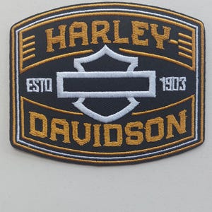 May include: A rectangular embroidered patch with rounded corners featuring a black background, gold borders, and white accents. The text reads HARLEY-DAVIDSON with ESTD 1903 on the sides, centered around a white shield-shaped emblem graphic.