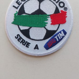 May include: Circular patch with a football design, the text reads "LEGA CALCIO SERIE A". The patch has green, white, and red accents, and a blue rectangle with the word "TIM".