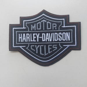 Può includere: Una toppa ricamata nera a forma di scudo con la scritta MOTOR HARLEY-DAVIDSON CYCLES in filo bianco e grigio. Il design presenta un sottile bordo marrone e un contorno bianco, creando un emblema classico per capi di abbigliamento.