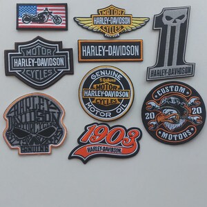 Könnte beinhalten: Eine Sammlung von acht gestickten Harley-Davidson-Aufnähern in verschiedenen Formen und Farben. Die Motive umfassen ein Motorrad auf einer US-Flagge, das Harley-Davidson-Logo, einen Totenkopf, die Zahl 1, ein 1903-Emblem und ein Adler-Grafikmotiv.