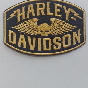 May include: An oval-shaped embroidered patch featuring a black background with gold-colored thread. The text reads HARLEY DAVIDSON in a bold font, accompanied by a central skull graphic with wings and two small lightning bolt symbols on the sides.