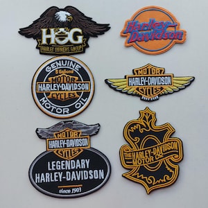 May include: Six embroidered patches featuring Harley-Davidson branding. Designs include a HOG eagle emblem, an orange circular logo, a round motor oil patch, a winged bar and shield, an oval legendary patch, and a decorative gold-bordered crest.