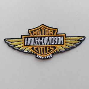 May include: An embroidered patch featuring the Harley-Davidson logo with a central shield shape, yellow wings extending to the sides, and black borders. The text reads MOTOR HARLEY-DAVIDSON CYCLES in white and yellow lettering on a black background.
