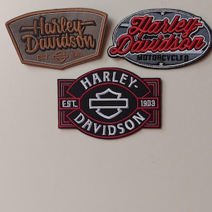 May include: Three embroidered Harley-Davidson patches. The top left is a brown trapezoid with black text. The top right is an oval with red text and grey accents. The bottom is a black rectangular patch with white text and a central shield logo.