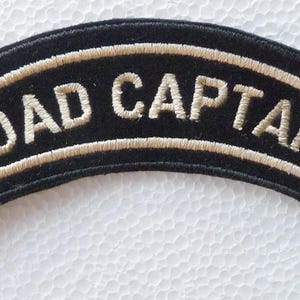 Puede incluir: Un parche bordado negro en forma de arco con el texto ROAD CAPTAIN en letras mayúsculas de color claro. El parche tiene un borde grueso de color claro que perfila la forma arqueada, creando un contraste marcado sobre el tejido negro.