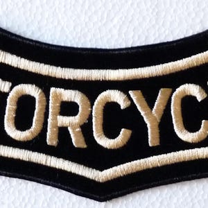 May include: A curved, black embroidered patch featuring the word MOTORCYCLES in bold, off-white capital letters. The patch has a distinct arched shape with a pointed bottom center and a thick, off-white stitched border outlining the entire design.