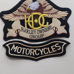 Könnte beinhalten: Zwei gestickte Aufnäher mit schwarzem Hintergrund. Der obere Aufnäher zeigt einen Weißkopfseeadler mit ausgebreiteten Flügeln über dem gelben Schriftzug HOG und den Worten HARLEY OWNERS GROUP auf einem Banner. Der untere Aufnäher ist ein schwarzes Banner mit dem Wort MOTORCYCLES.