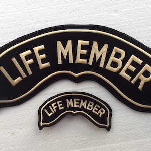 Könnte beinhalten: Zwei schwarze, gebogene Stickaufnäher, die jeweils den in Goldfaden gestickten Text "LIFE MEMBER" tragen. Der größere Aufnäher ist über dem kleineren positioniert, beide zeigen identische Schriftzüge und einen goldenen Rand.