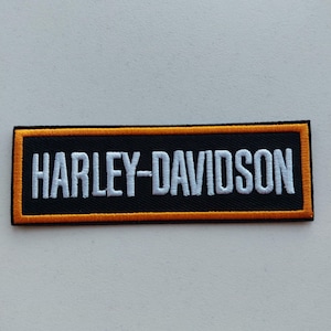 May include: A rectangular embroidered patch featuring the text HARLEY-DAVIDSON in white capital letters. The patch has a black background and is bordered by a thick orange frame, with a thin black outer edge surrounding the entire rectangular shape.