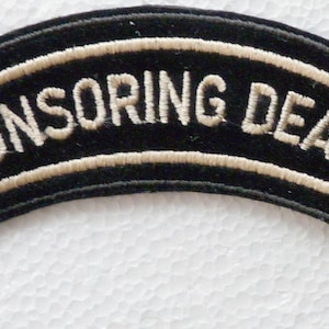 Puede incluir: Un parche bordado negro de forma curva que presenta el texto "SPONSORING DEALER" en letras mayúsculas blancas. El parche tiene un borde blanco que delinea el texto y el borde exterior de la forma curva, creando un contraste marcado con la tela negra.