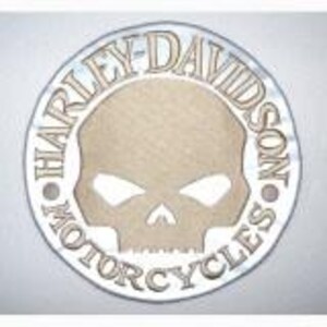 Könnte beinhalten: Ein runder Aufnäher mit einer beigen Totenkopf-Grafik, die von einem weißen Rand umgeben ist. Der Schriftzug HARLEY-DAVIDSON MOTORCYCLES ist in beiger Schrift entlang des kreisförmigen Randes um das Totenkopf-Design gedruckt.