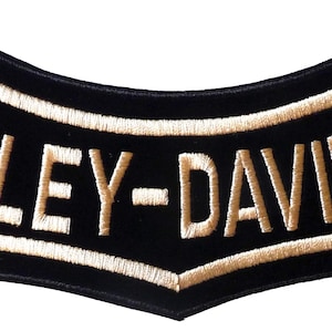 May include: A curved, black embroidered patch featuring the text HARLEY-DAVIDSON in bold, tan-colored thread. The patch has a distinct arched shape with a tan border outlining the lettering and the outer edge of the black fabric base.