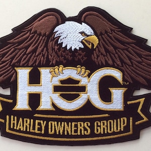 May include: An embroidered patch featuring a brown eagle with a white head and yellow beak, positioned above the white text HOG and the gold text HARLEY OWNERS GROUP on a black background. The design includes a shield graphic within the letter O.