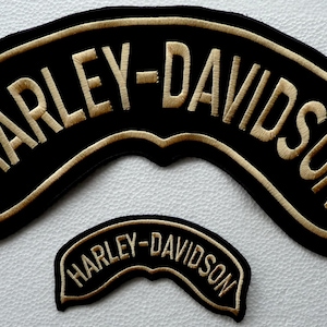 Può includere: Due toppe in tessuto nero a forma di arco con bordi ricamati in oro e il testo "HARLEY-DAVIDSON" in lettere dorate. La toppa più grande è posizionata sopra quella più piccola, entrambe presentano un bordo inferiore curvo.