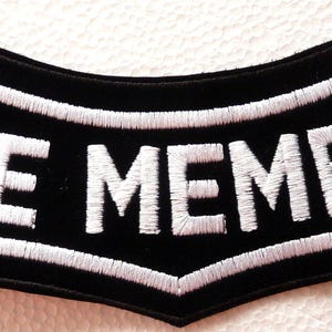 May include: A black embroidered patch with a curved, banner-like shape featuring the white text LIFE MEMBER in a bold, sans-serif font. The patch has a thick white stitched border that follows the contour of the black fabric background.