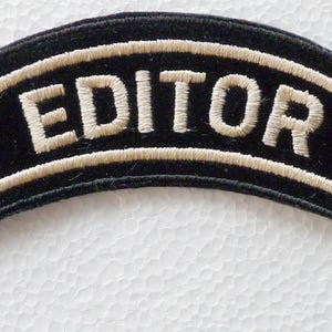 Puede incluir: Un parche bordado negro en forma de arco con la palabra EDITOR en letras mayúsculas de color blanco hueso. El parche tiene un borde grueso cosido en color blanco hueso que sigue la forma curva, creando un diseño de alto contraste.