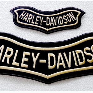 Op de afbeelding: Twee zwarte geborduurde patches in een gebogen, bannerachtige vorm. Elke patch bevat de tekst HARLEY-DAVIDSON gestikt in een licht crèmekleurige of gebroken witte draad, met een bijpassende rand op de zwarte stoffen achtergrond.