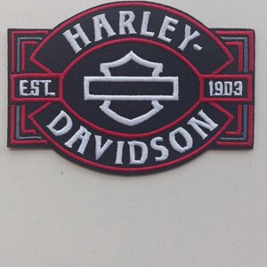 Op de afbeelding: Een rechthoekige geborduurde patch met een zwarte achtergrond en rood-witte randen. De tekst HARLEY-DAVIDSON is in het wit geborduurd, met EST. 1903 aan de zijkanten. Een wit schildembleem is gecentreerd in het ovale ontwerp.