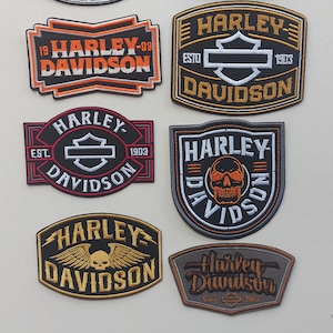 Könnte beinhalten: Eine Sammlung von Harley-Davidson-Patches in verschiedenen Formen und Farben. Die Patches zeigen das Harley-Davidson-Logo, Text und Grafiken, darunter ein Schädel- und Flügeldesign. Einige Patches enthalten das Jahr 1903.