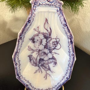 May include: A white ceramic shell-shaped dish featuring a deep purple floral transferware pattern in the center and a decorative border. The dish has a scalloped edge and is displayed on a metal stand, showcasing intricate botanical details.