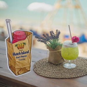 May include: A sticker of a brown paper bag with a red bottle and straw, with the text "HI from Hutch Island Fort Pierce Florida". A green and white drink with a straw and pink decoration sits on a woven coaster. A small lavender plant in a burlap sack is also present.
