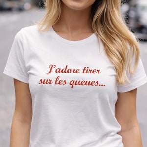 Peut inclure: T-shirt blanc avec l'inscription en script rouge "J'adore tirer sur les queues...". Le t-shirt est associé à un jean en denim bleu. Le modèle sourit, avec des cheveux blonds.