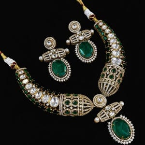 May include: A luxury jewellery set comprising a necklace and matching earrings. The necklace and earrings are decorated with emerald-green oval gemstones, surrounded by sparkling clear stones. The set has a gold-coloured metal setting against a black background.
