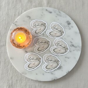 May include: A round marble tray displays oyster-shaped stickers, each bearing the name "Savannah". A small, lit candle in a peach-toned glass holder is on the left. The stickers have a pearl-like centre. The tray is on a neutral-toned surface.