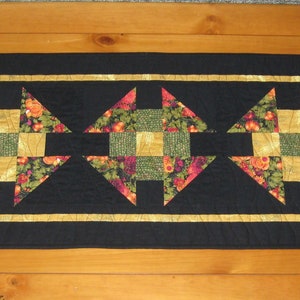 May include: A wooden coffee table with a black quilted table runner featuring a geometric pattern of gold, green, and orange fabric squares.