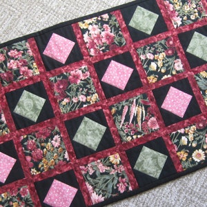 May include: A black, red, and green quilted table runner with a diamond pattern. The runner features floral fabric with pink, white, and green flowers.