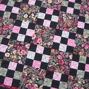 May include: A quilt with a checkered pattern in black, white, and pink. The quilt is made of fabric with a floral print in shades of pink, green, and brown.