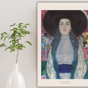 Puede incluir: Una impresión enmarcada de la pintura de Gustav Klimt, que representa a una mujer con un gran sombrero negro y un vestido azul y blanco. El fondo tiene pinceladas coloridas de rosa, verde y amarillo. Un jarrón blanco con una planta verde está en primer plano.