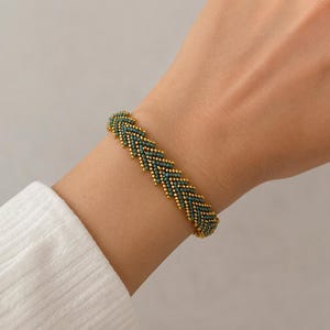 Puede incluir: Una pulsera de cuentas con un patrón de espiga repetitivo construido con pequeñas cuentas de oro metálico y cuentas de color verde azulado oscuro. La pulsera tiene una apariencia tejida y estructurada con una textura consistente en toda la banda.