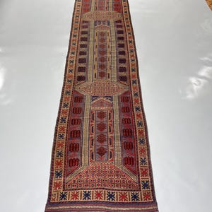 Könnte beinhalten: Ein langer, schmaler, rechteckiger Teppich mit einem detaillierten geometrischen Muster. Der Teppich hat ein zentrales Design in Rot-, Braun- und Beigetönen mit blauen und anderen farbigen Bordüren. Der Teppich ist wahrscheinlich aus gewebtem Material.