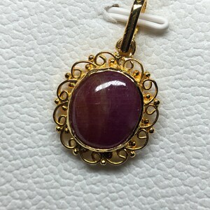 May include: A gold pendant with an oval, deep red gemstone. The gemstone is set in a decorative gold filigree frame. The pendant has a gold bail for attaching to a chain. The background is a textured white surface.