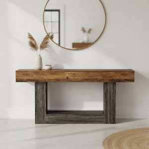 May include: A wooden console table with a rectangular top and two gray, rectangular legs. The table is made of wood with a natural finish. A gold-framed mirror hangs on the white wall above the table. A vase with dried plants and a small bowl sit on the table.